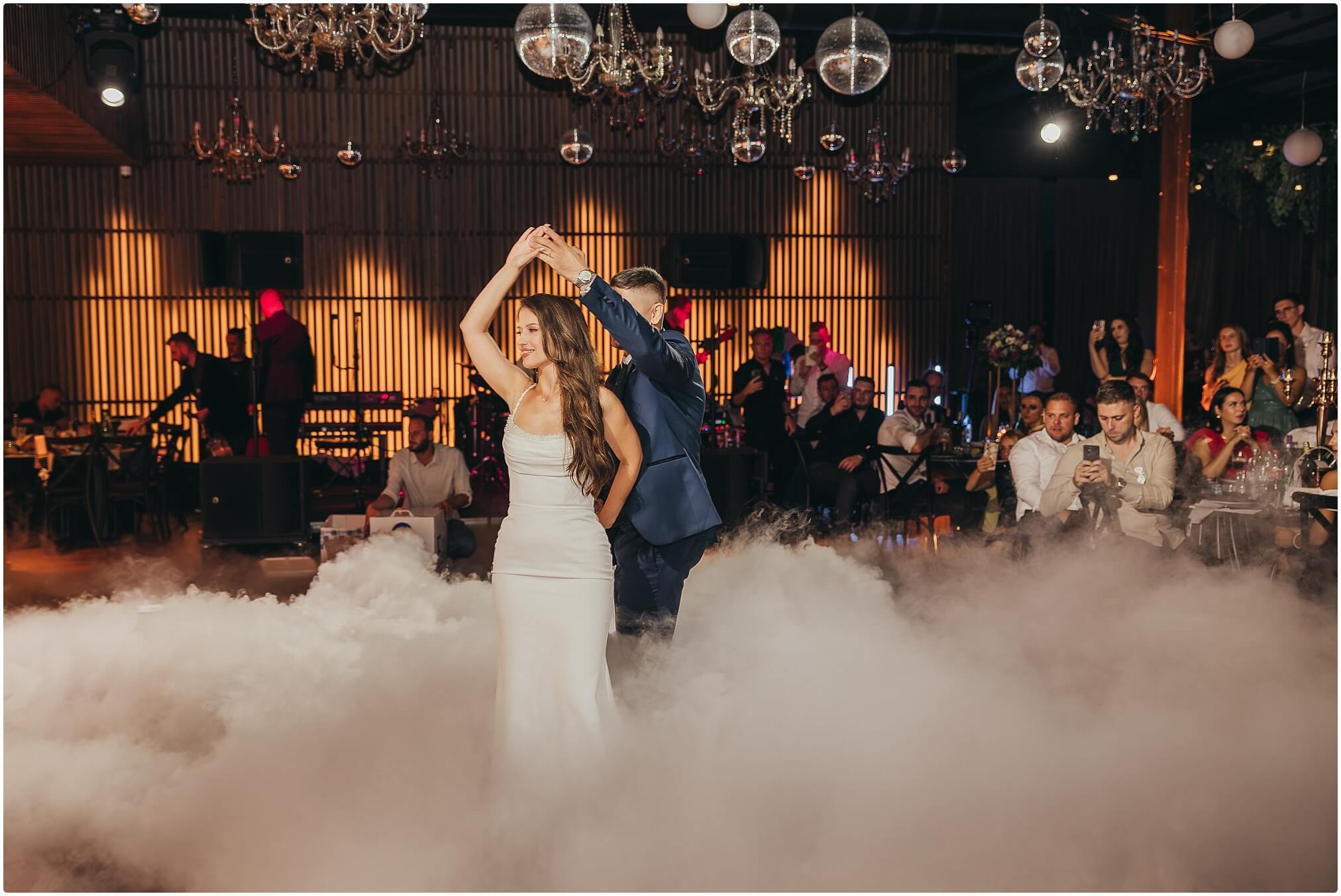 Mladenci plešu prvi ples u elegantnoj dvorani, okruženi prijateljima i svjetlima